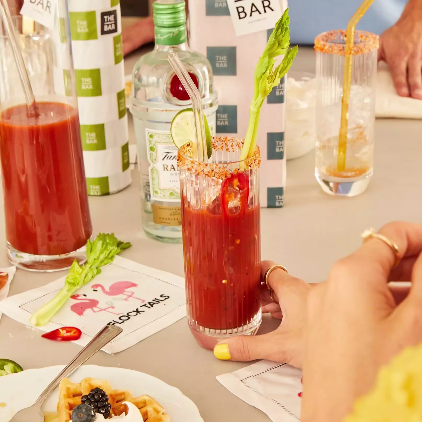 A hand holds a Bloody Mary garnished with celery and chili; other drinks, a gin bottle, and napkins with flamingos are on the table.