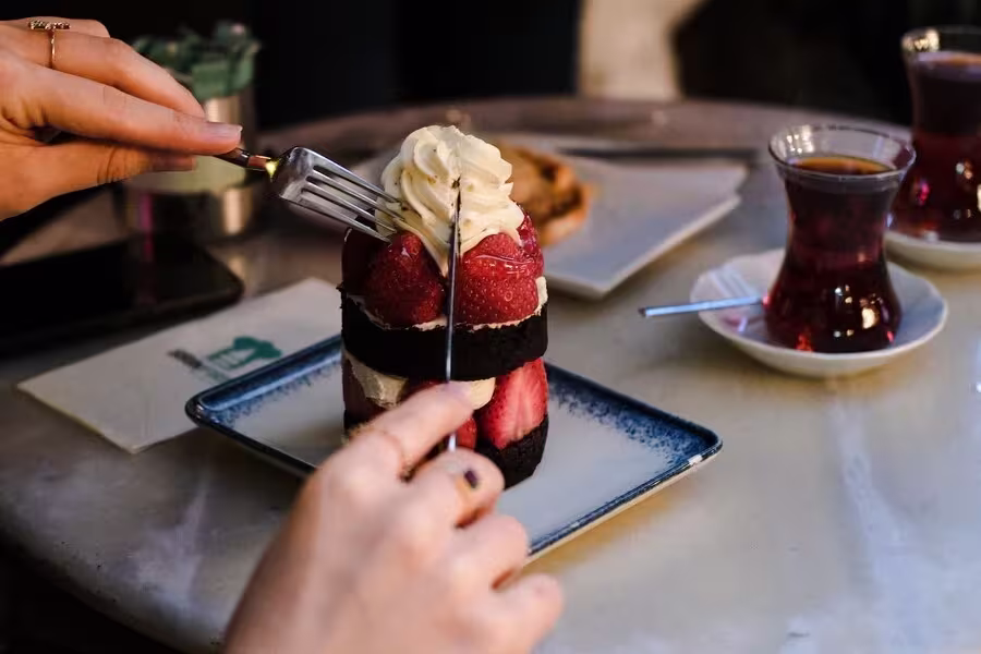 A person cuts into a layered chocolate and strawberry dessert with cream, while two glasses of tea and a napkin rest on the table.
