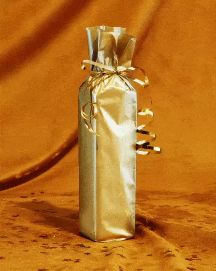 A bottle wrapped in shiny gold paper with gold ribbon, standing upright against a matching gold fabric background.