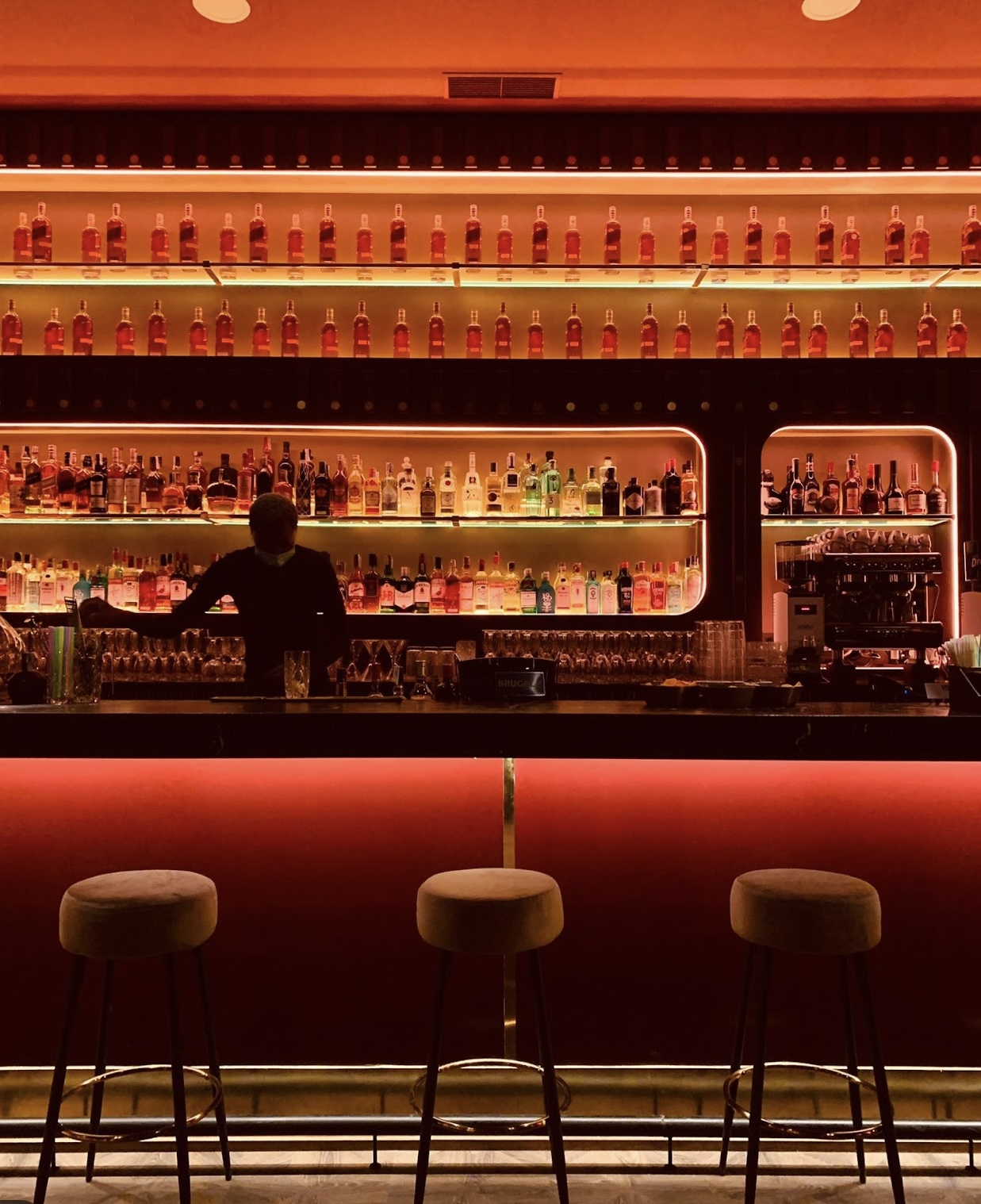 La imagen muestra el interior de la Coctelería Montana, con una elegante barra iluminada en tonos cálidos y estantes llenos de botellas perfectamente alineadas. El ambiente es sofisticado y acogedor.