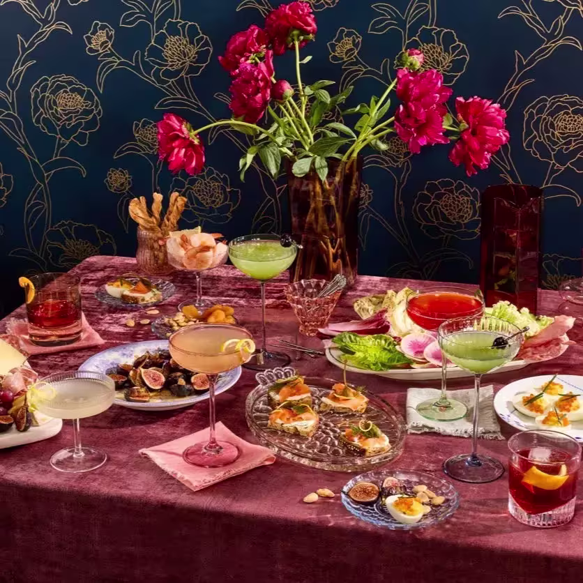 A table set with various cocktails, appetizers, and small plates, decorated with a vase of red flowers against a floral-patterned dark blue wall.