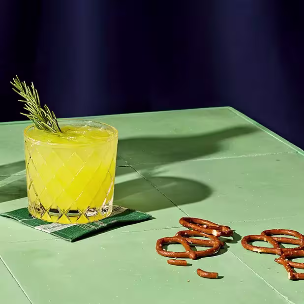 A glass of yellow cocktail garnished with a rosemary sprig sits on a green napkin atop a green table, with several pretzels scattered nearby.