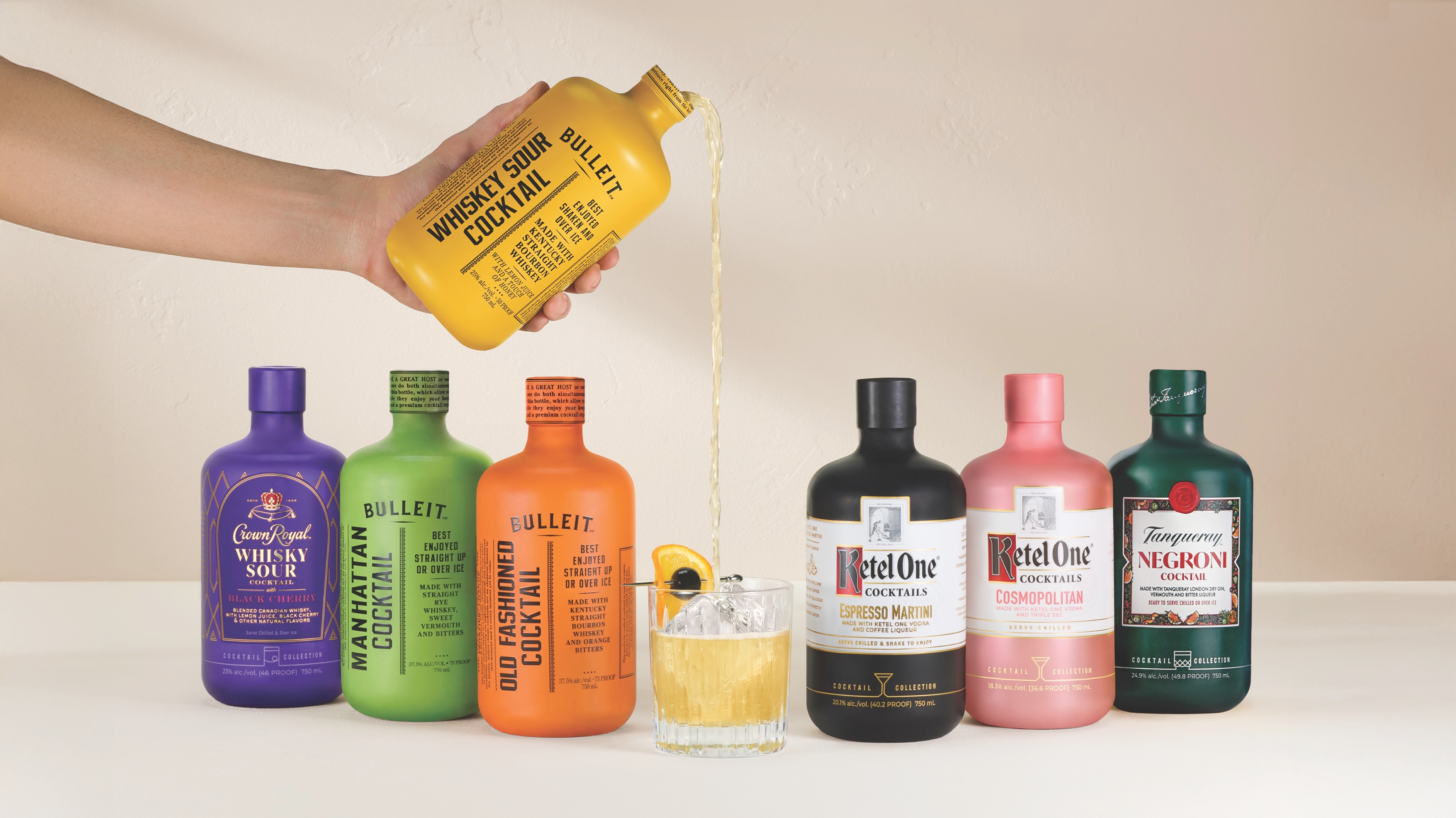 A hand pours Bulleit Whiskey Sour cocktail from a yellow bottle into a glass with ice and an orange slice, surrounded by six assorted bottled cocktails.