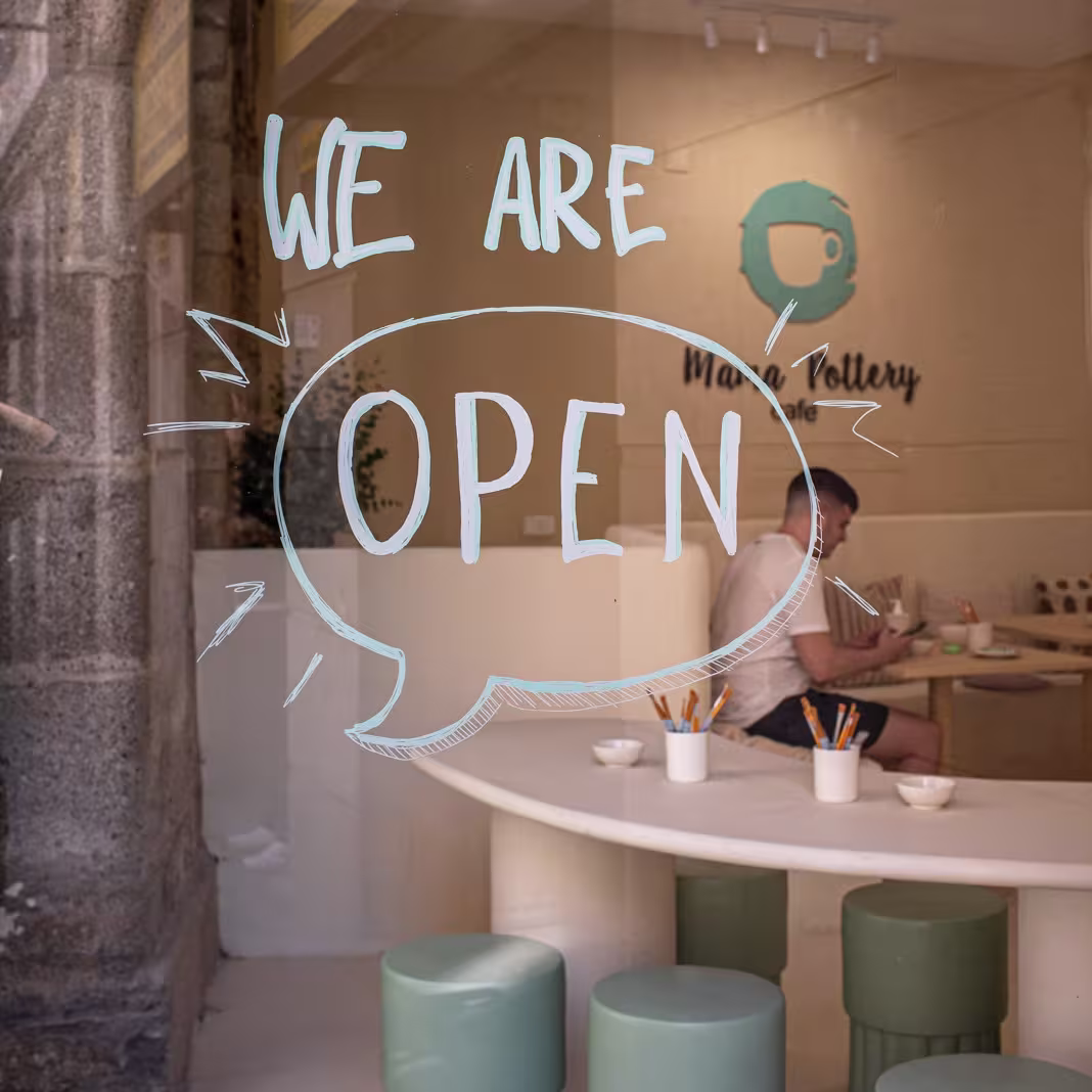Cristalera de Mama Pottery Café con el mensaje “We are open” escrito a mano, interior luminoso con bancos redondos y una persona disfrutando del espacio.