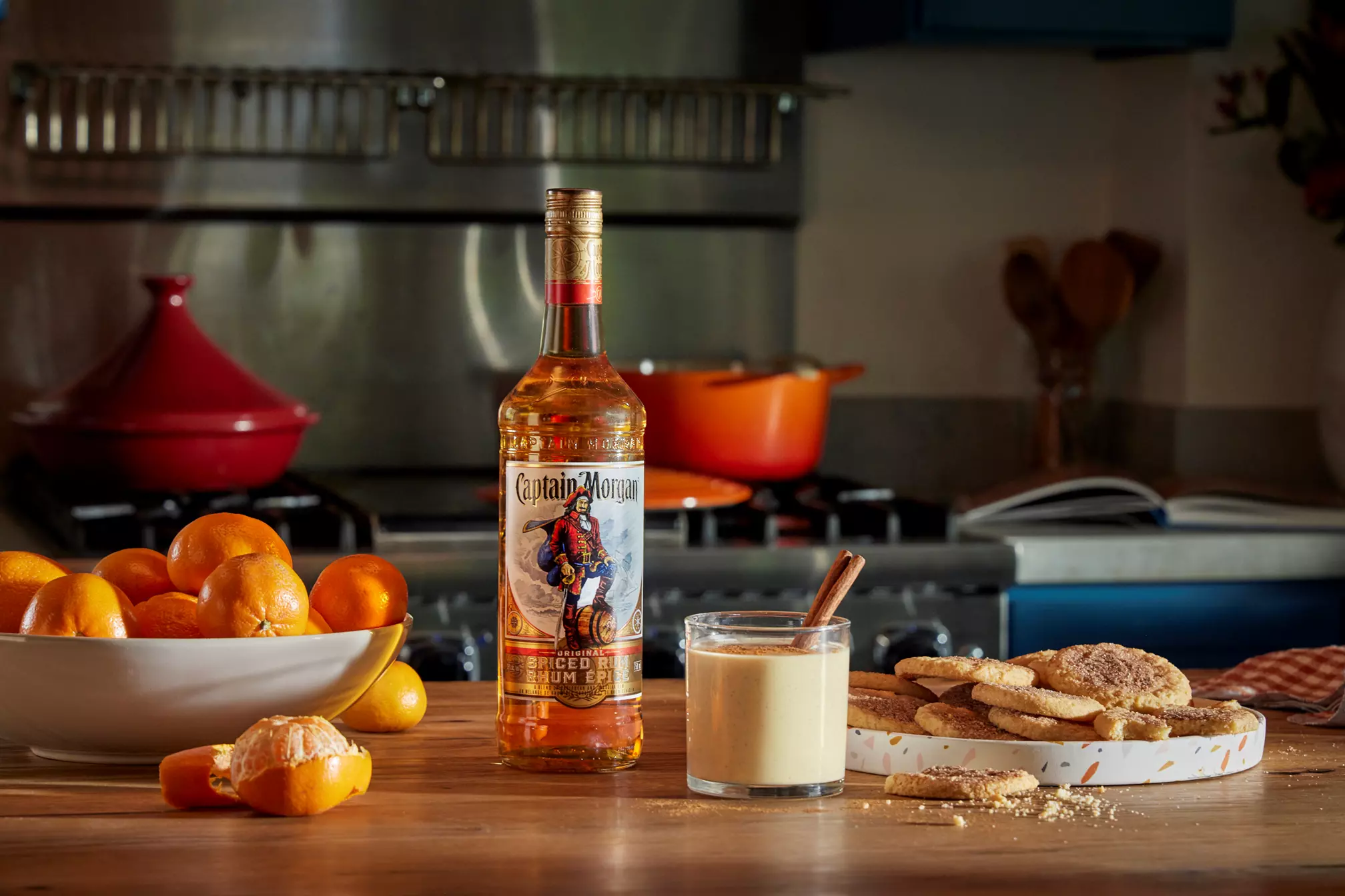 A bottle of Captain Morgan spiced rum, a glass of eggnog with a cinnamon stick, a plate of cookies, and a bowl of oranges are arranged on a kitchen counter.