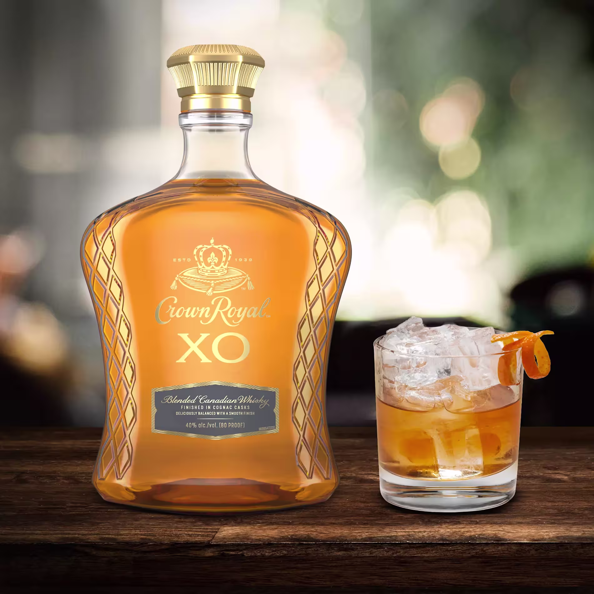 A bottle of Crown Royal XO blended Canadian whisky sits next to a glass of whisky with ice and an orange twist on a wooden surface.