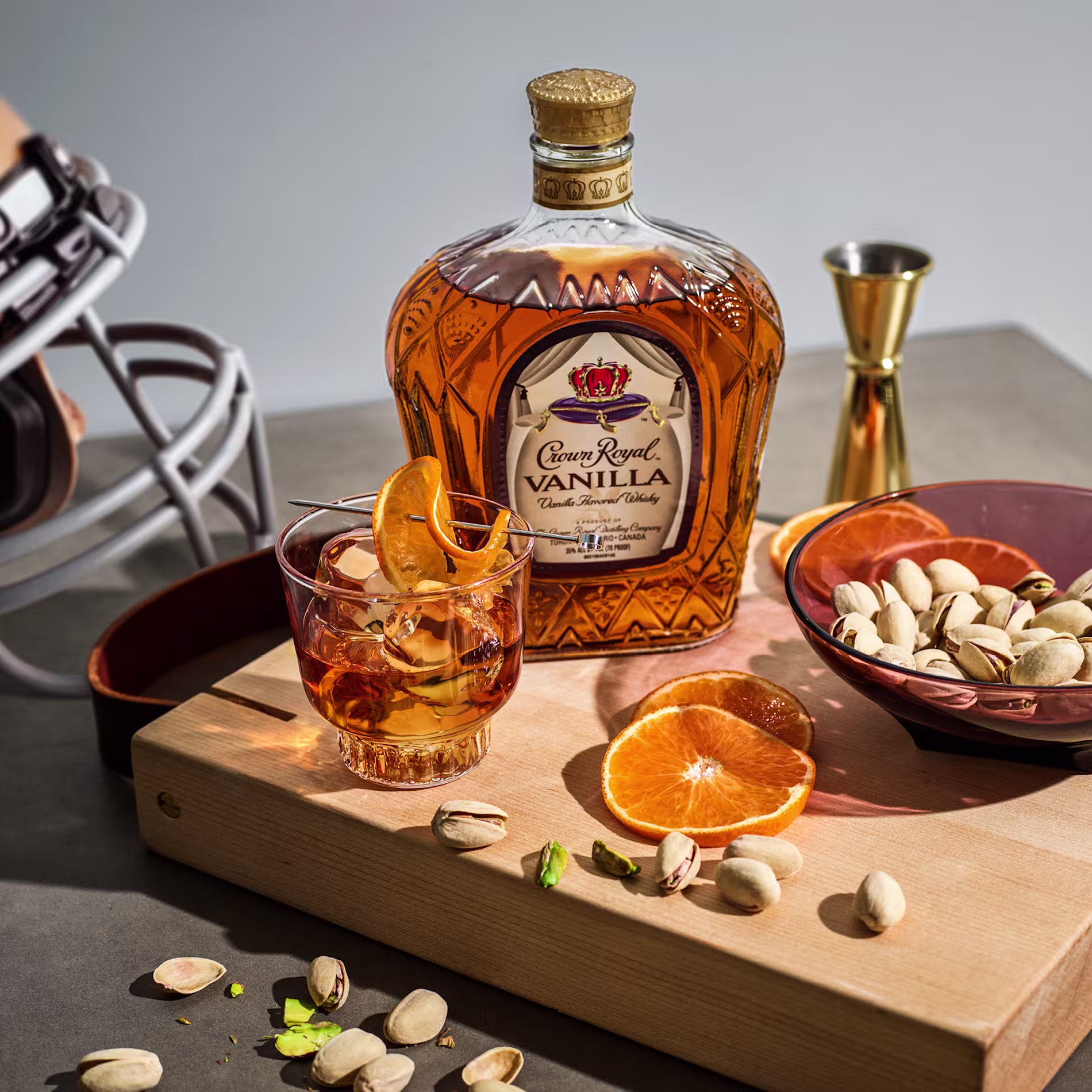 A bottle of Crown Royal Vanilla whiskey, a glass with whiskey and orange garnish, pistachios, a jigger, and orange slices on a wooden board, with a football helmet in the background.