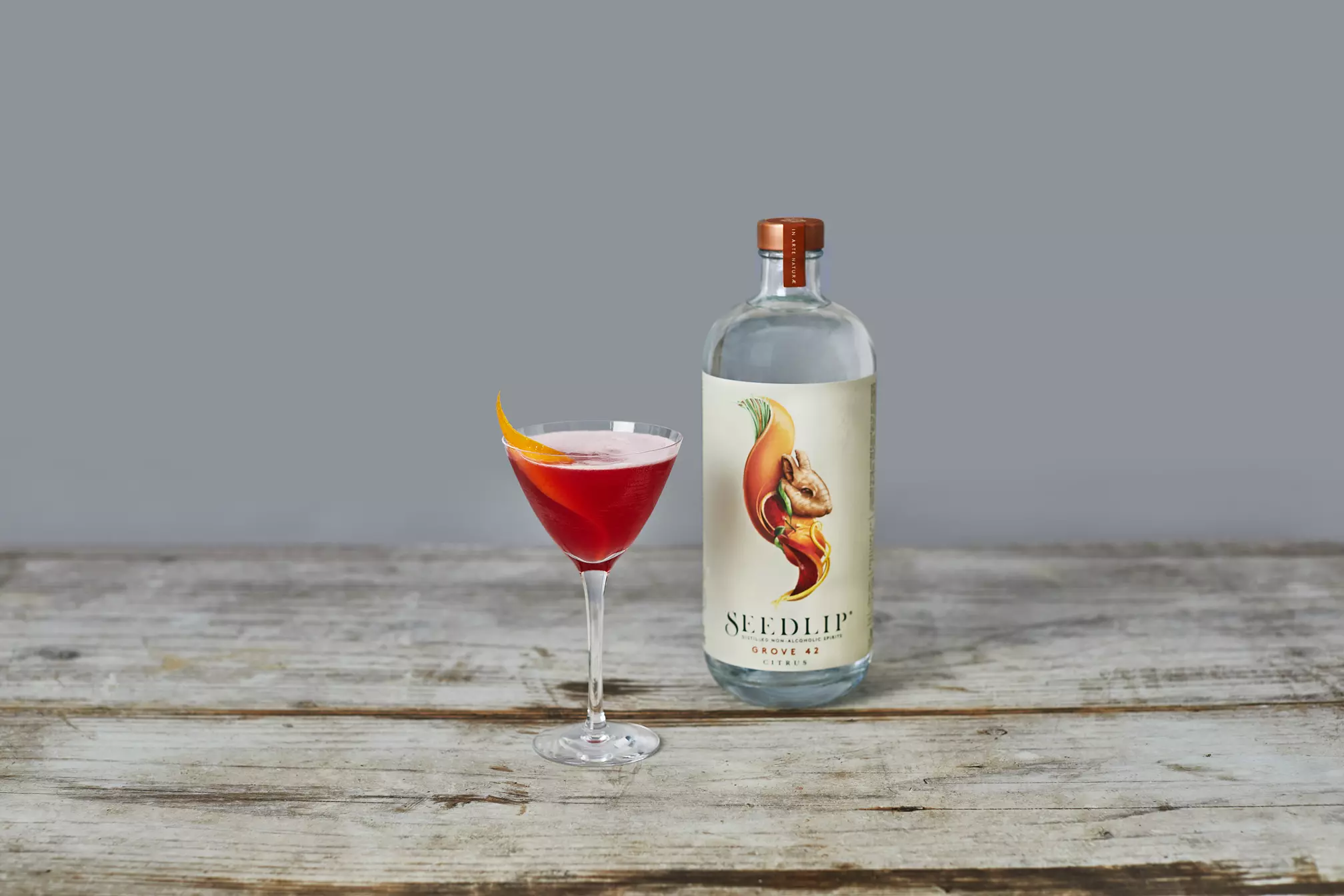 A bottle of Seedlip Grove 42 and a red cocktail in a coupe glass garnished with an orange twist, placed on a rustic wooden surface.