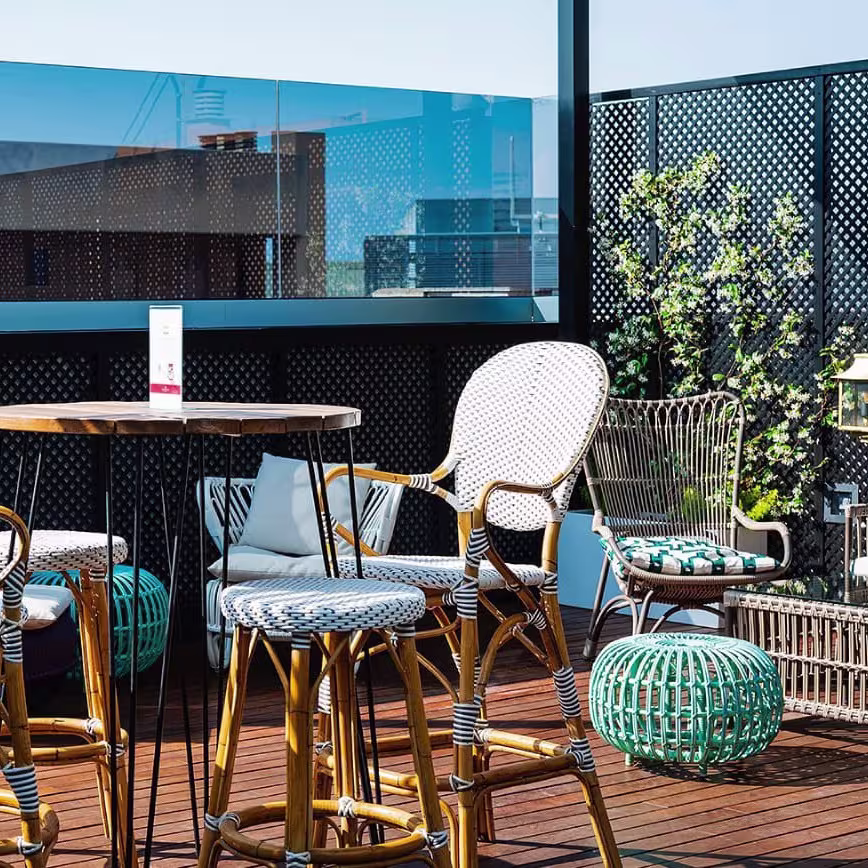 Terraza luminosa con mesas de madera, sillas de mimbre y detalles vegetales sobre suelo de madera, rodeada de celosías negras.