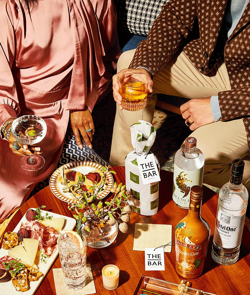Two people sit at a table with various drinks, liquor bottles, and appetizers, including charcuterie and olives. One person holds a cocktail, the other a glass of amber liquid.