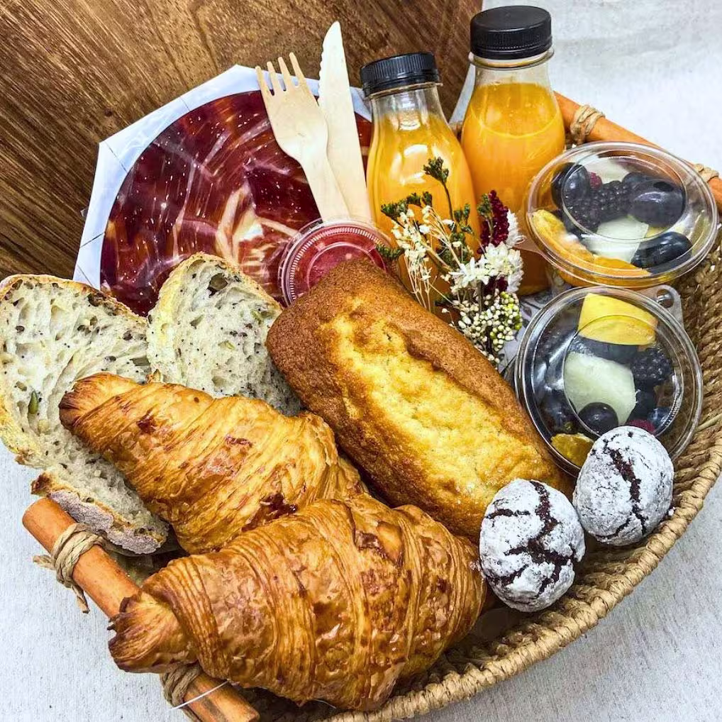 Cesta de desayuno con croissants, bizcocho, pan de semillas, fruta fresca, zumo de naranja, dulces y jamón en lonchas sobre fondo de madera.