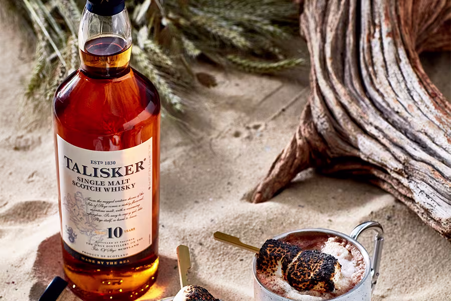 A bottle of Talisker 10-year-old single malt whisky stands on sand next to a mug topped with toasted marshmallows, with driftwood and dried plants in the background.