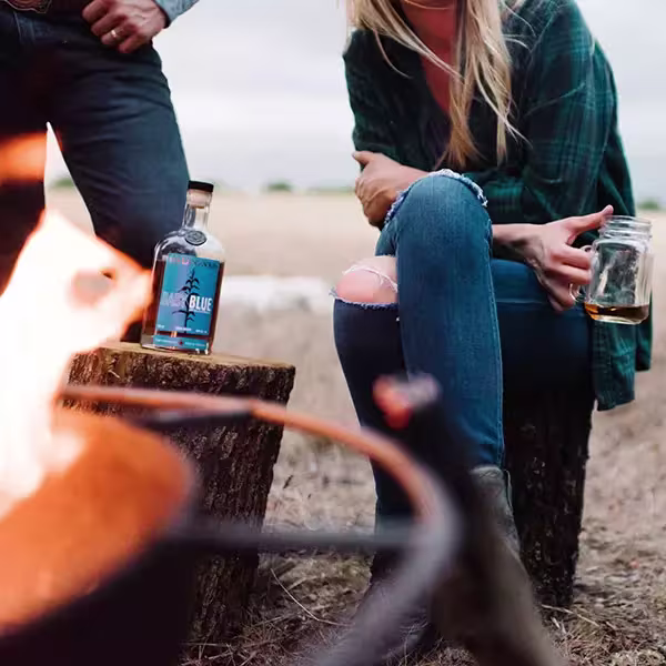 Two people sit on tree stumps outdoors near a fire pit; one holds a jar with a drink, and a bottle is placed on a stump.
