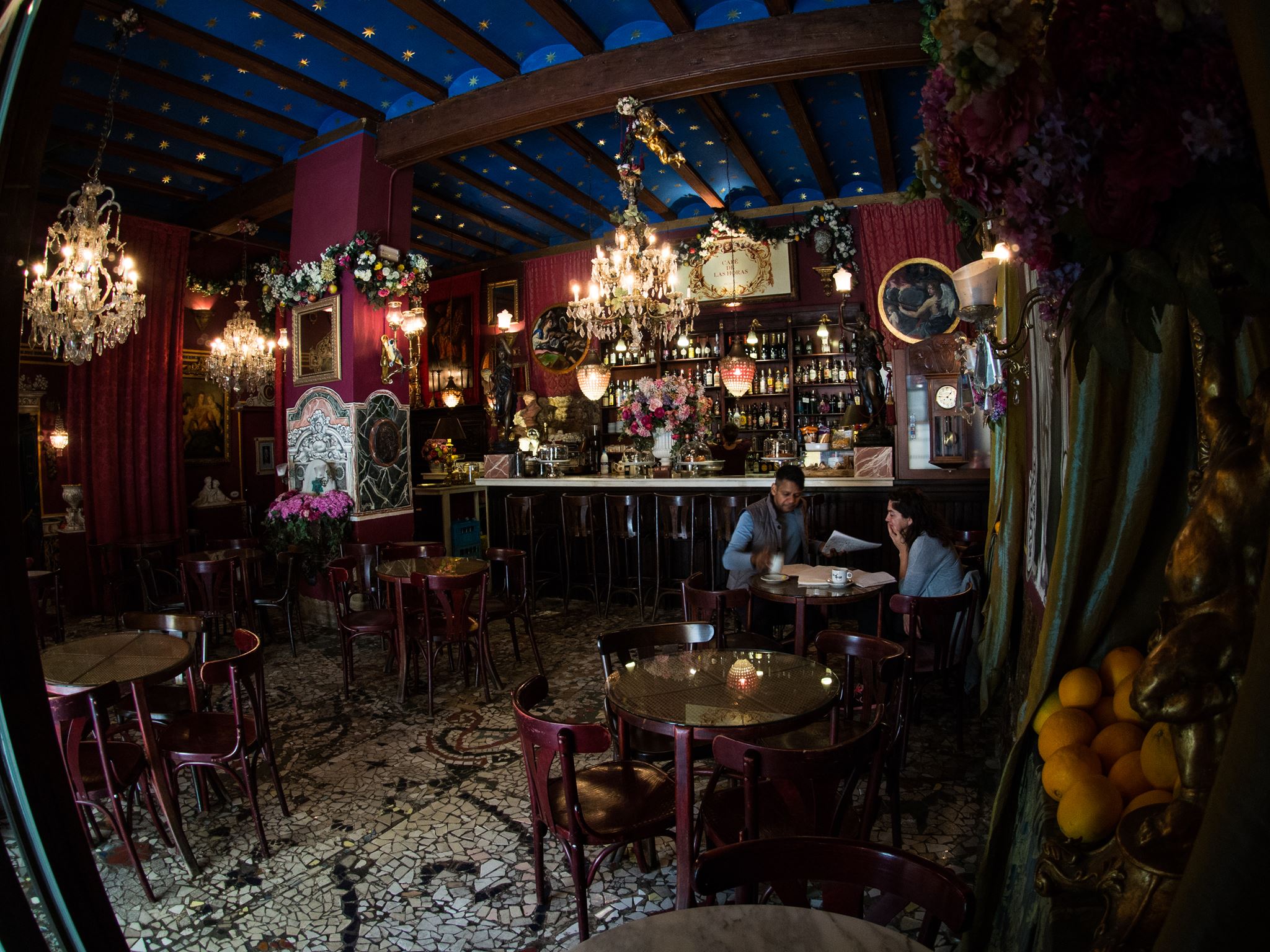 Interior de Café de las Horas con techo azul estrellado, lámparas de araña, decoración floral y una pareja tomando algo en un ambiente íntimo.