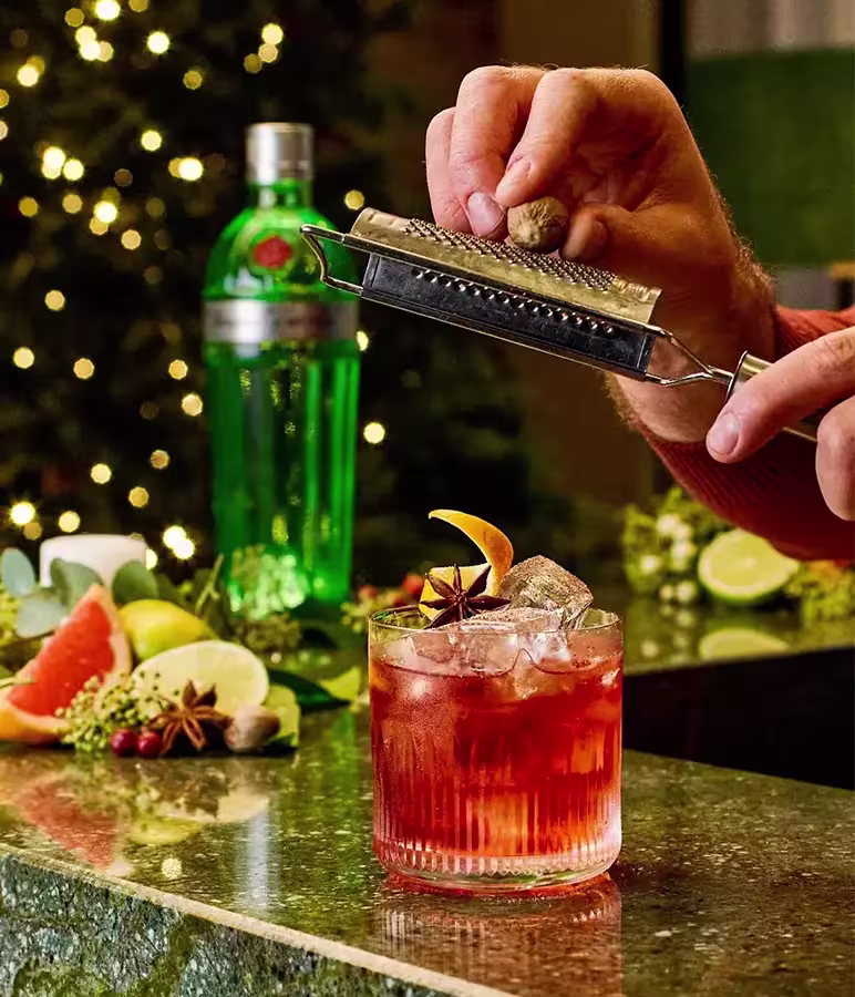 A hand grates nutmeg over a red cocktail with ice and citrus garnish on a bar, with a green bottle and festive decorations in the background.