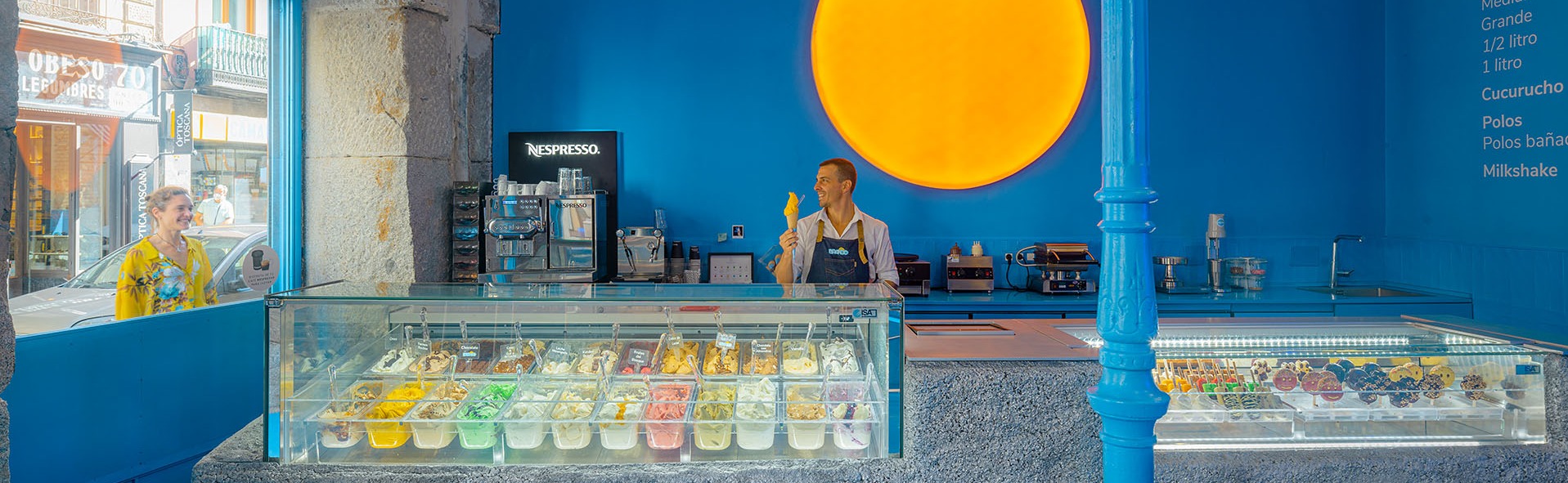 Heladería Brando con interior azul vibrante, gran sol luminoso en la pared, vitrinas repletas de helados de colores y polos decorados, atendida con una sonrisa.
