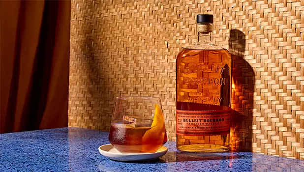 A bottle of Bulleit Bourbon whiskey stands next to a glass of whiskey with ice and an orange twist, placed on a blue surface against a woven background.