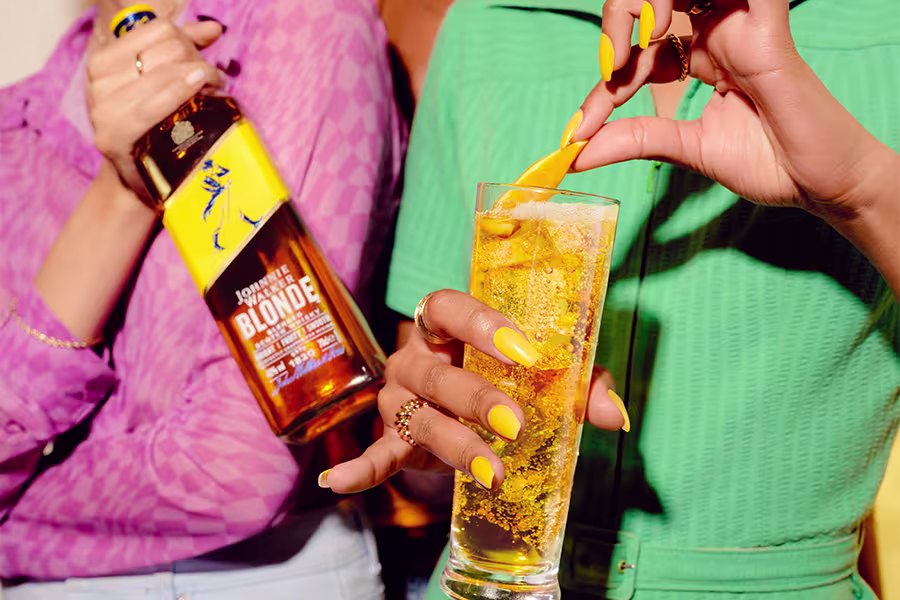 Two people, one holding a bottle of Johnnie Walker Blonde whisky, and the other garnishing a glass of whisky with an orange peel. Both have yellow manicured nails.