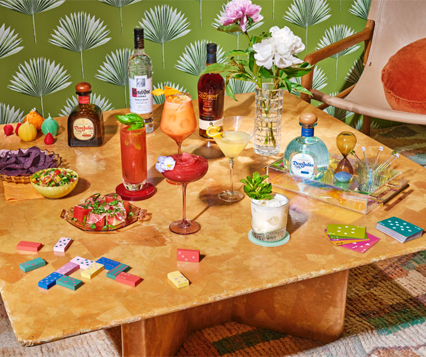 A table set with various cocktails, liquor bottles, snacks, a vase of flowers, dominos, and playing cards, against a green patterned wallpaper background.