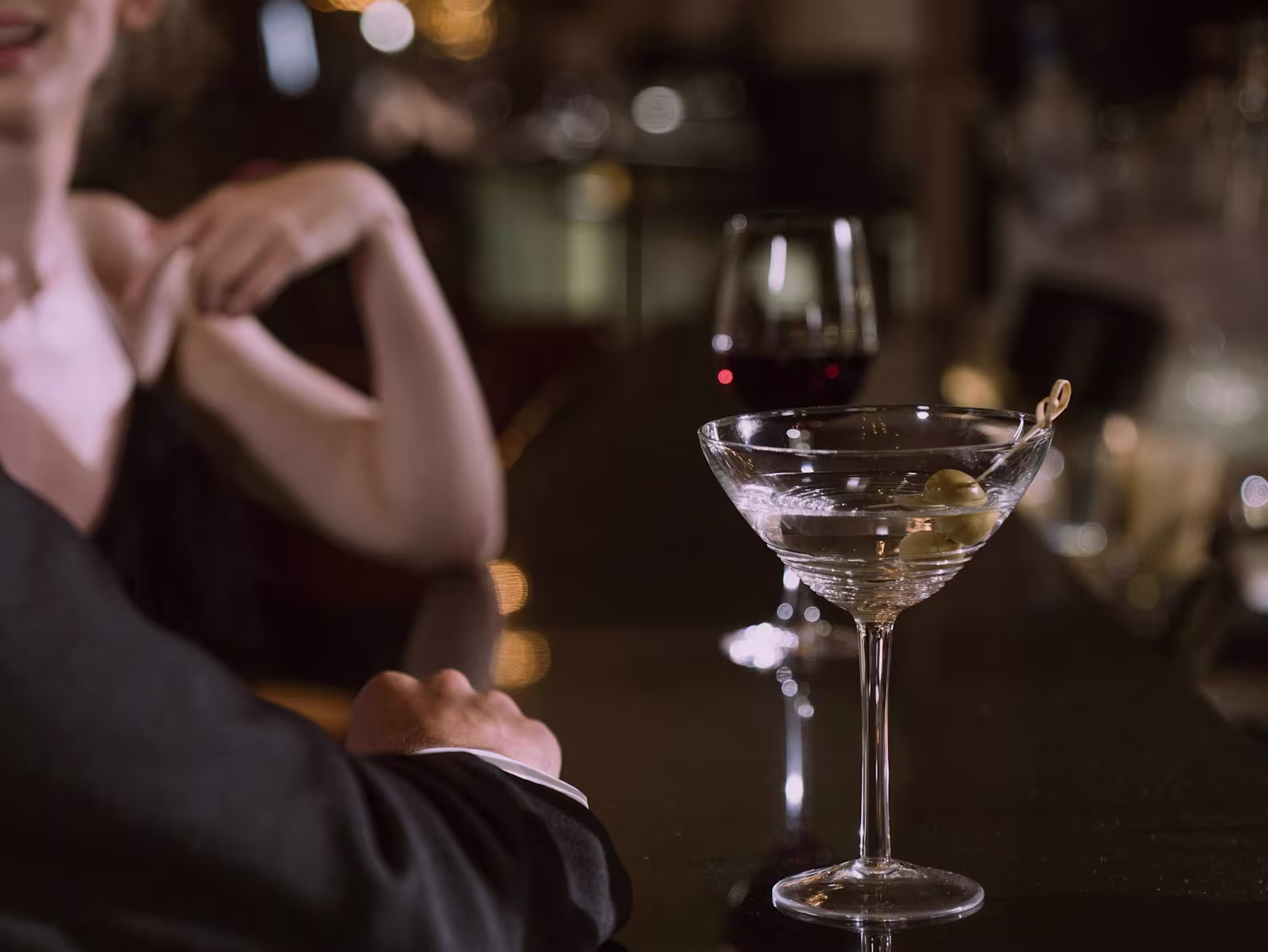 A martini with an olive and a glass of red wine sit on a bar counter; two people in formal attire are partially visible in the background.