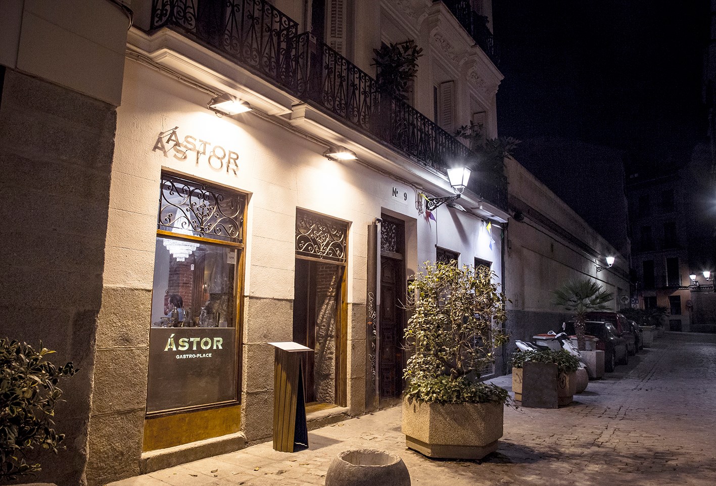 Fachada de Ástor Gastro-Place de noche, con iluminación cálida, ventanas decoradas y una calle tranquila con plantas y farolas.