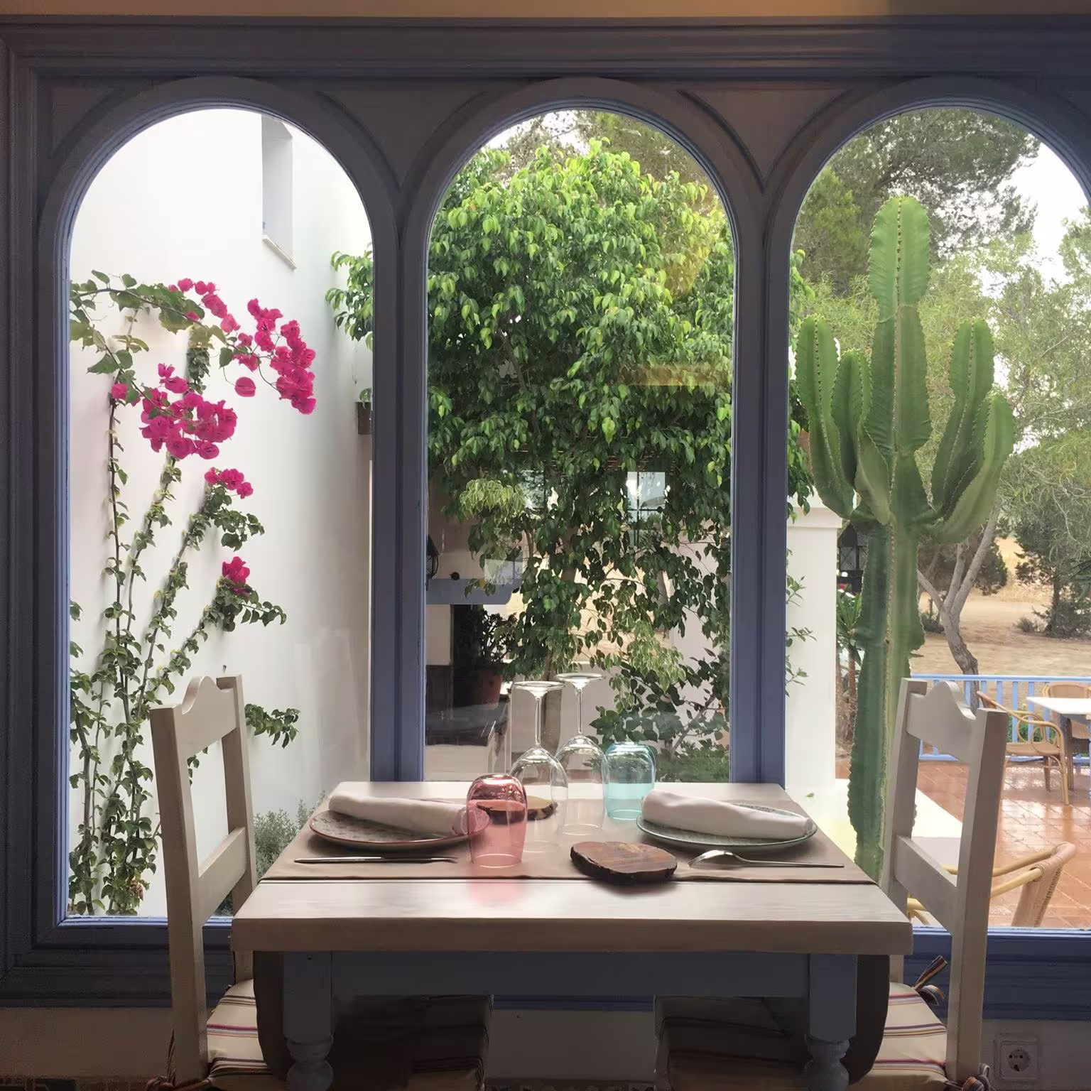 La imagen muestra la tranquila atmósfera de Es Tragon, con una mesa preparada junto a una ventana que enmarca un jardín lleno de flores y vegetación