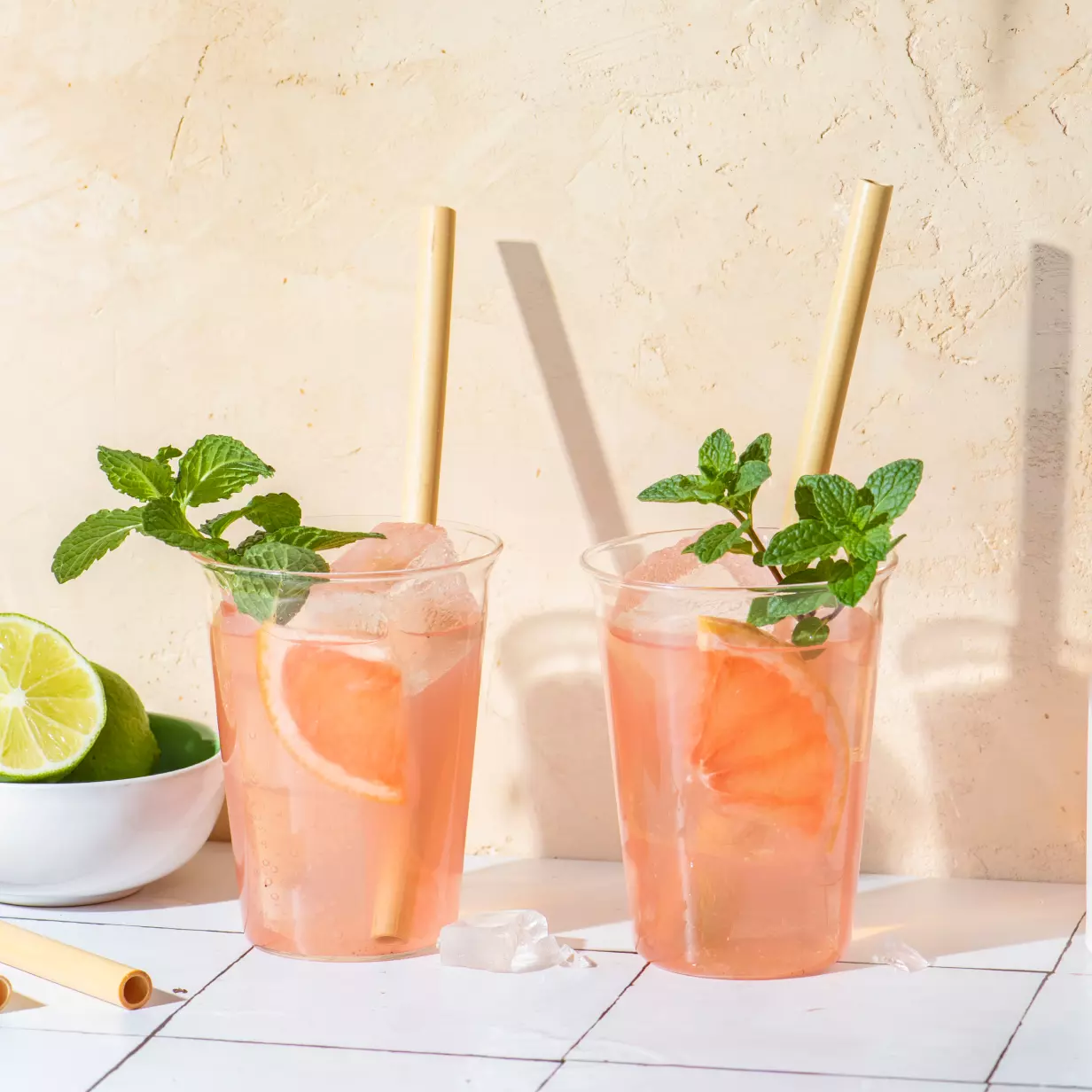 Two plastic cups with pink drinks, ice cubes, grapefruit slices, mint sprigs, and bamboo straws on a tiled surface; a bowl of limes is in the background.