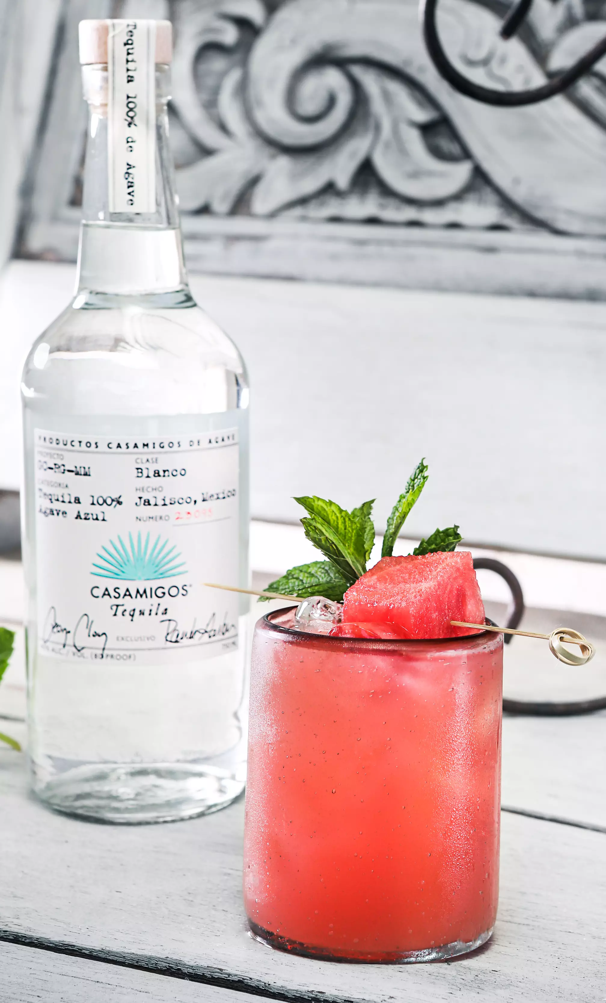 A glass of pink watermelon cocktail with ice and mint garnish sits in front of a bottle of Casamigos Blanco Tequila on a white wooden surface.