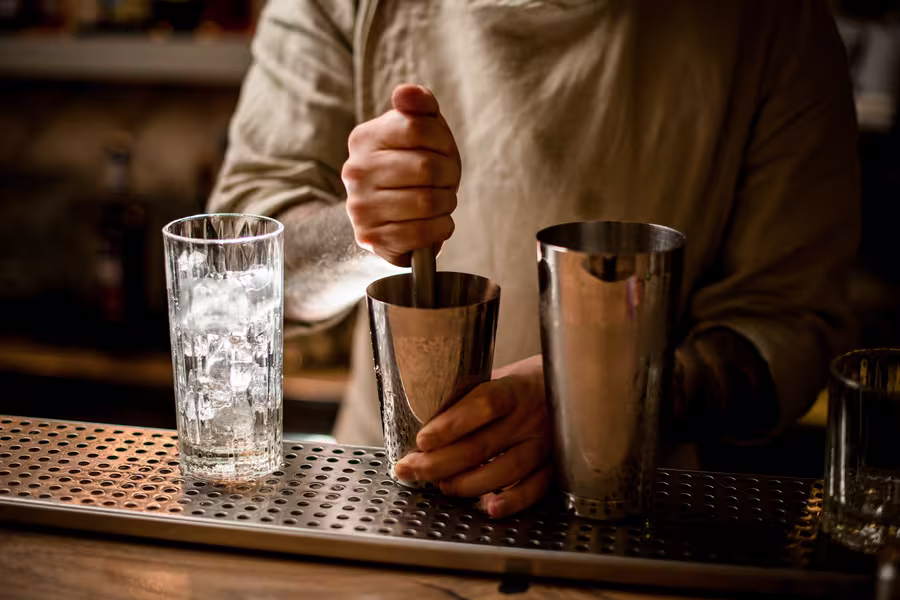 A person uses a muddler in a cocktail shaker beside a glass filled with ice on a bar counter.