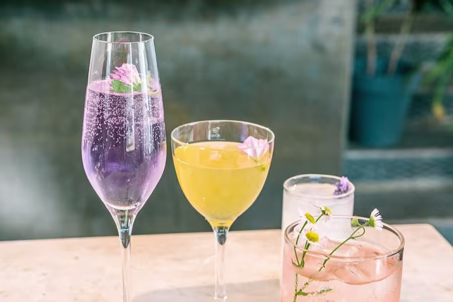 Four colorful cocktails in different glasses, each garnished with edible flowers, are arranged on a light-colored surface with a blurred background.