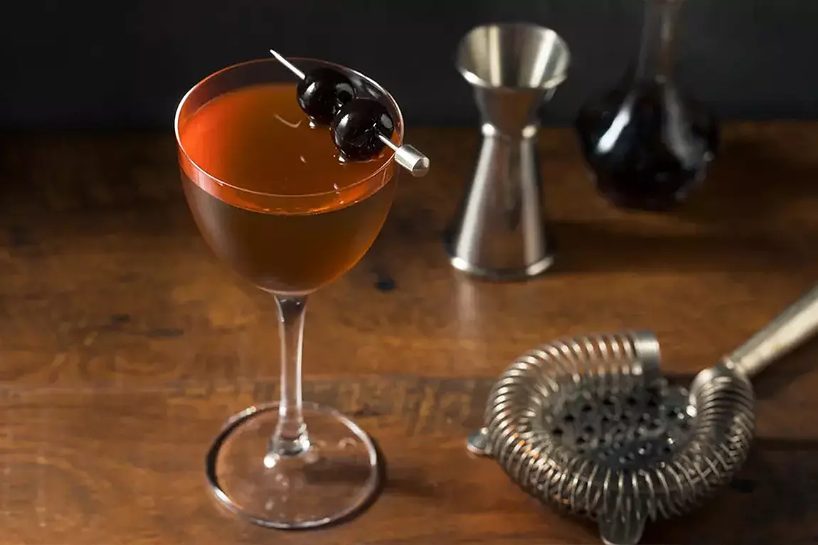 A Manhattan cocktail in a stemmed glass garnished with two dark cherries on a pick, placed on a wooden surface with a strainer, jigger, and bottle in the background.