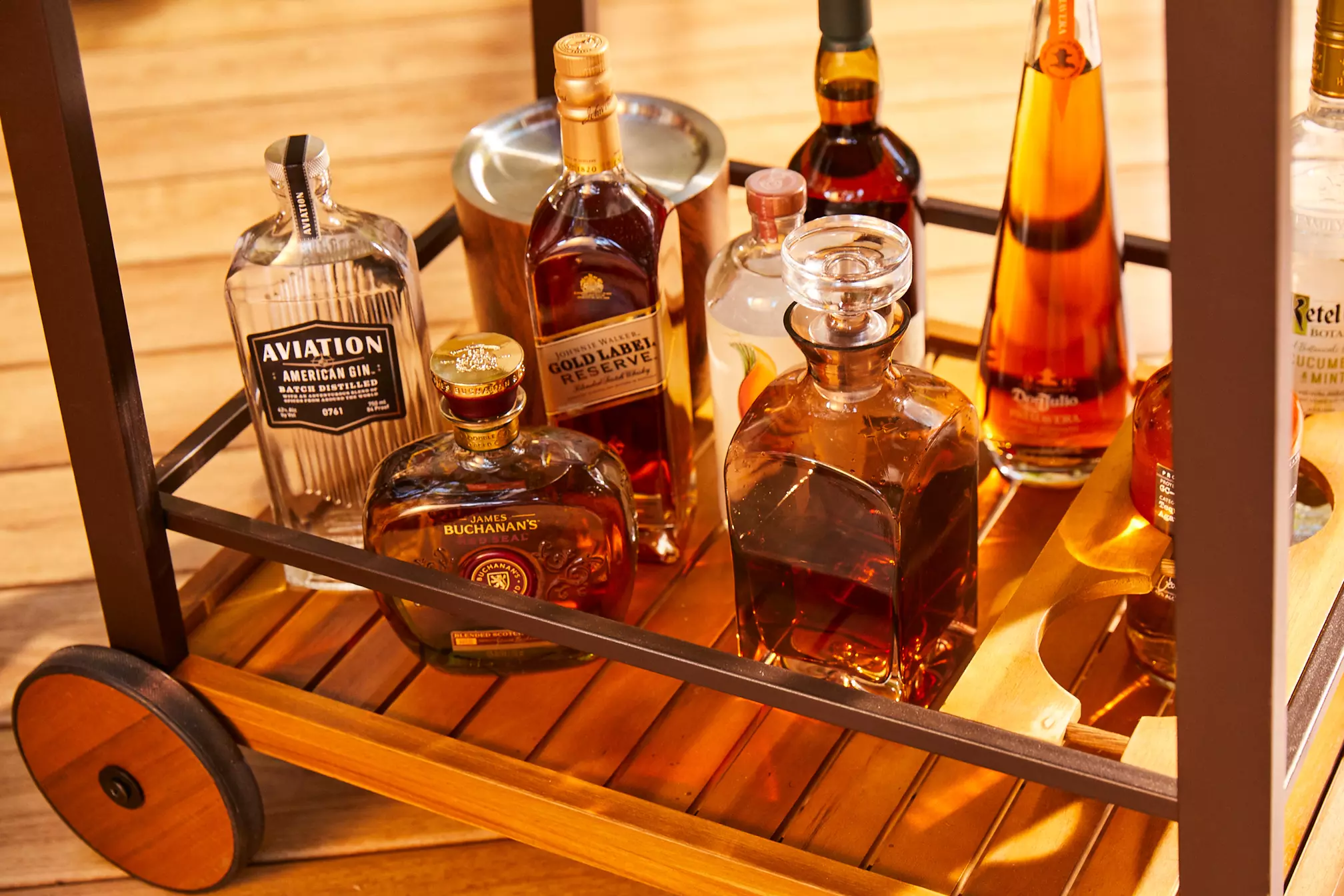 A wooden bar cart holding several bottles of liquor, including whiskey, gin, and decanters, placed on a wooden floor.