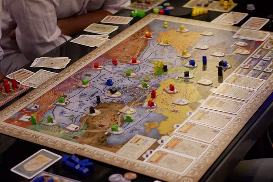A board game in progress with colored pieces, cards, and tokens on a map-themed game board surrounded by players.