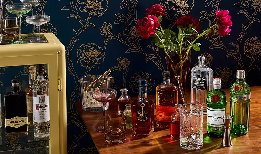 A wooden bar counter displays various bottles of liquor, cocktail glasses, a mixing glass, and a vase with red flowers against a floral-patterned dark blue wall.
