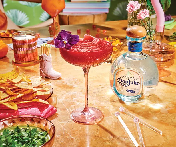 A pink frozen cocktail in a glass with a purple flower garnish sits on a table with Don Julio tequila, chips, dips, flowers, and decorative items.