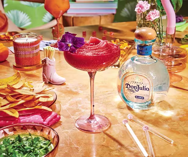 A pink frozen cocktail in a glass with a purple flower garnish sits on a table with Don Julio tequila, chips, dips, flowers, and decorative items.