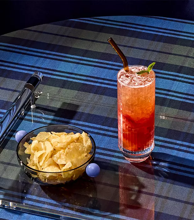 A glass of red iced drink with a straw and mint garnish sits next to a bowl of potato chips on a glass table with a blue plaid tablecloth.