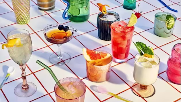 A variety of colorful cocktails and drinks in different glasses are arranged on a white table with a red grid pattern.
