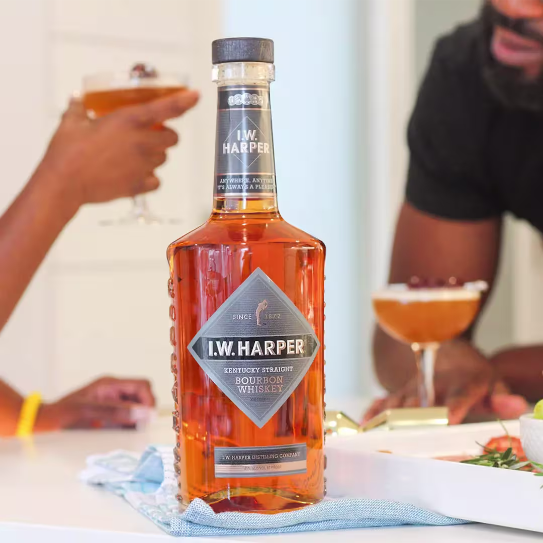 A bottle of I.W. Harper Kentucky Straight Bourbon Whiskey is placed on a table, with two people holding cocktails in the background.