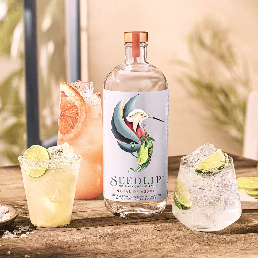 A bottle of Seedlip non-alcoholic spirit sits on a wooden table surrounded by three cocktails garnished with lime and grapefruit slices.