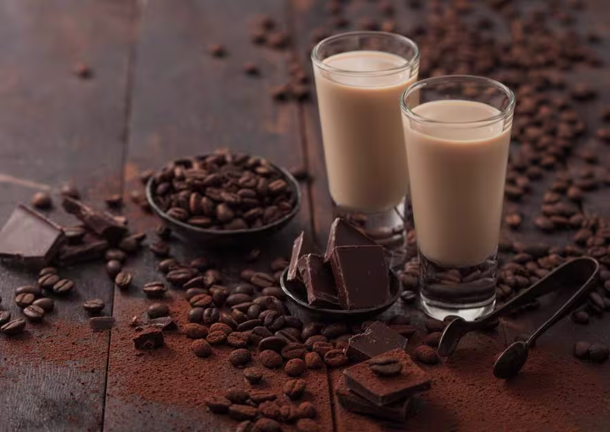 Two glasses of coffee liqueur sit on a wooden table surrounded by coffee beans, chocolate pieces, and cocoa powder.