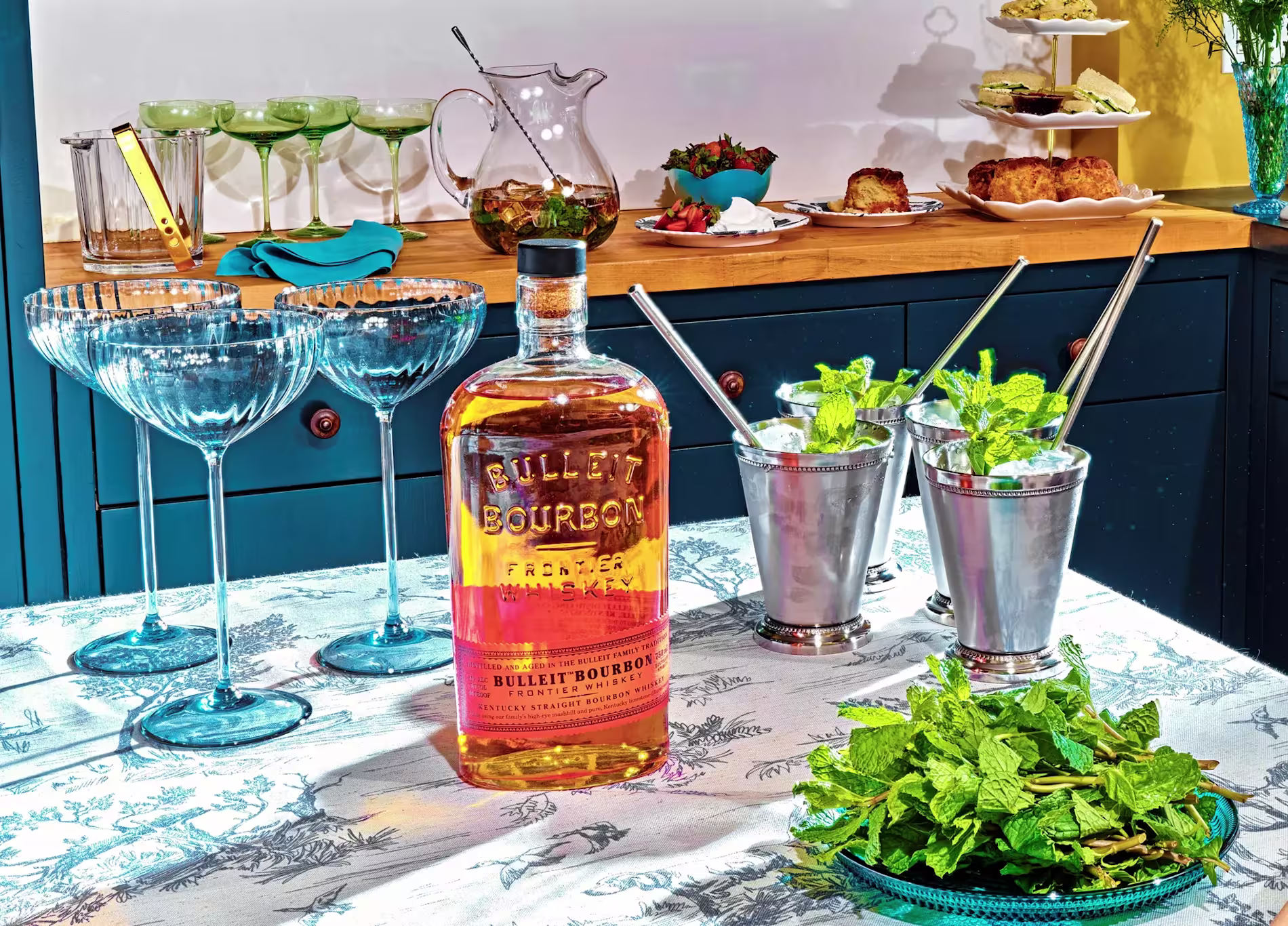 A bottle of Bulleit Bourbon, empty cocktail glasses, mint juleps, fresh mint, and assorted food are arranged on a table and counter for a gathering.