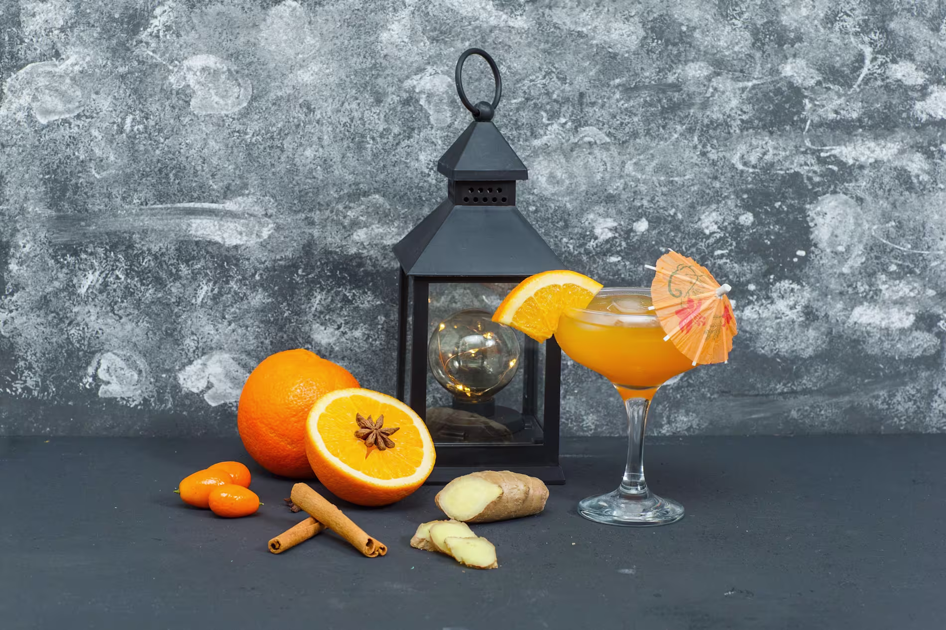 A glass of orange cocktail with an umbrella, garnished with an orange slice, sits beside fresh oranges, kumquats, cinnamon sticks, ginger, and a black lantern against a gray background.