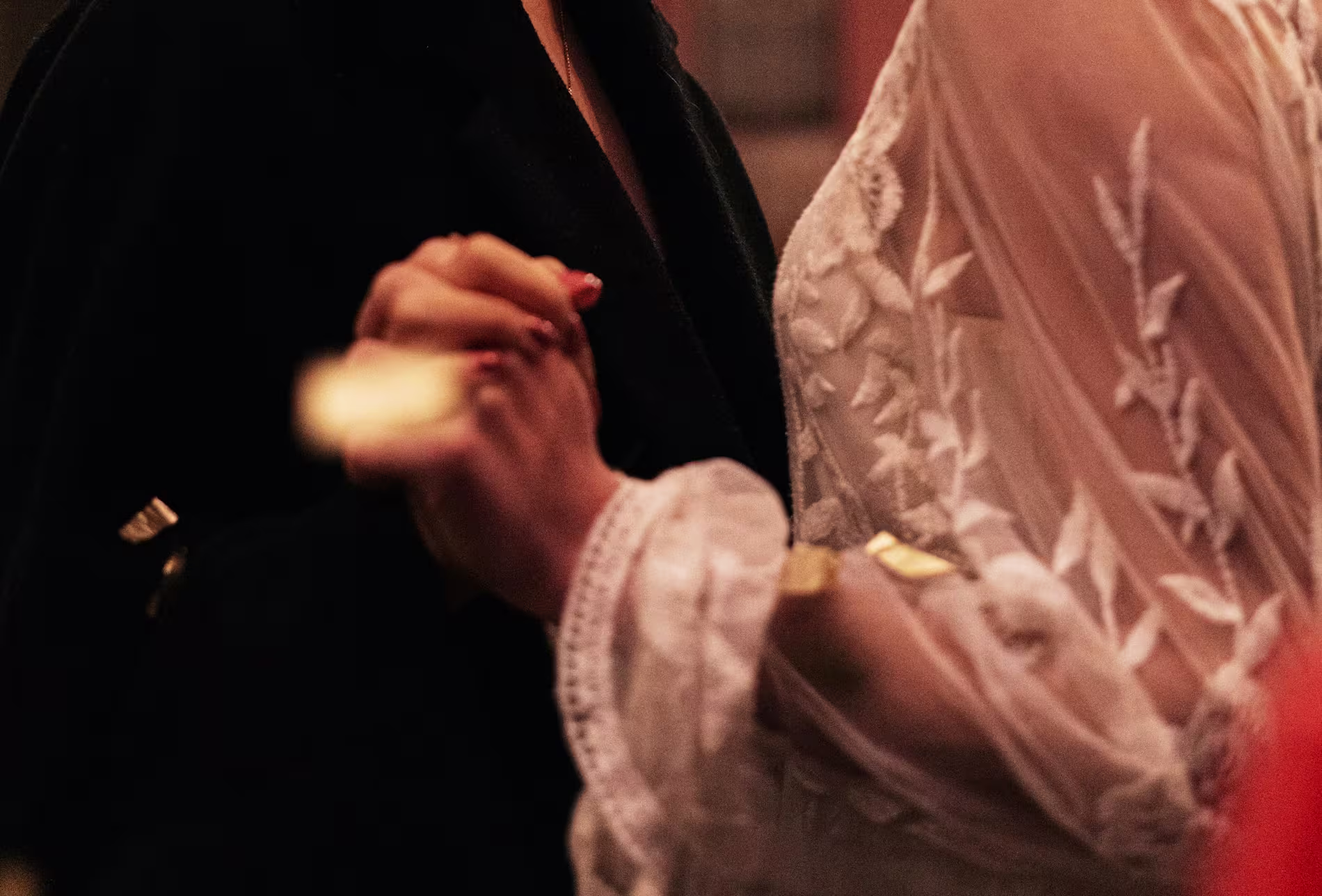 Two people are holding hands; one is wearing a black garment, and the other is wearing a white lace outfit with embroidered details.