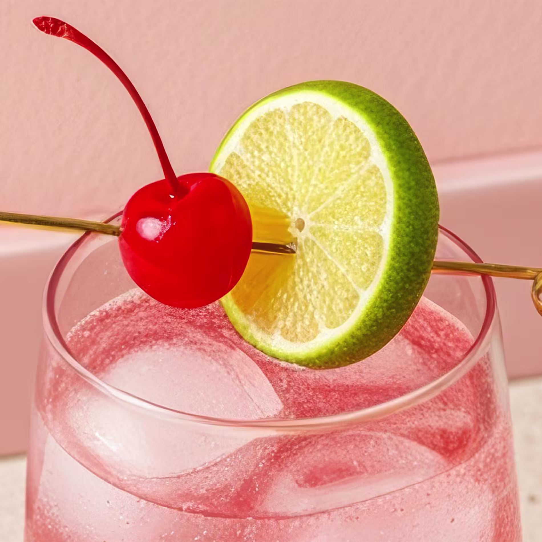 A lime wheel and a cherry on a metal skewered, laid across the rim of a glass with a pink cocktail