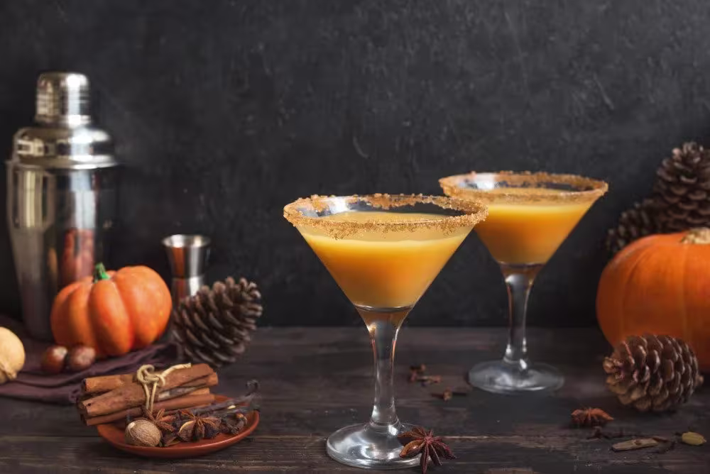 Two pumpkin-colored cocktails in martini glasses with spiced rims on a rustic table, surrounded by pumpkins, pinecones, cinnamon sticks, and a cocktail shaker.