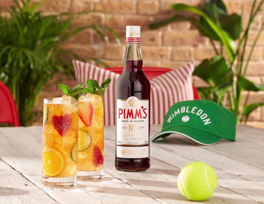 Pimm’s No.1 bottle with fruit-filled glasses, tennis ball, and Wimbledon cap on a table.