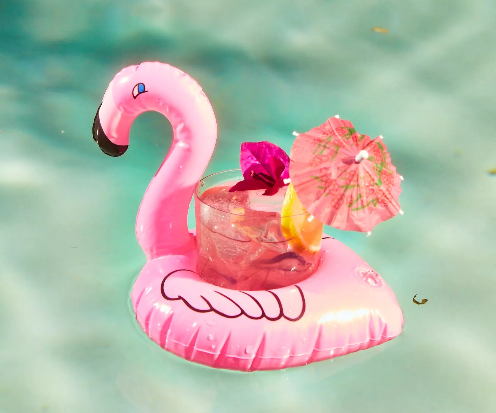 A pink inflatable flamingo cup holder floats in a pool, holding a drink with ice, a lemon slice, a pink cocktail umbrella, and a decorative stirrer.