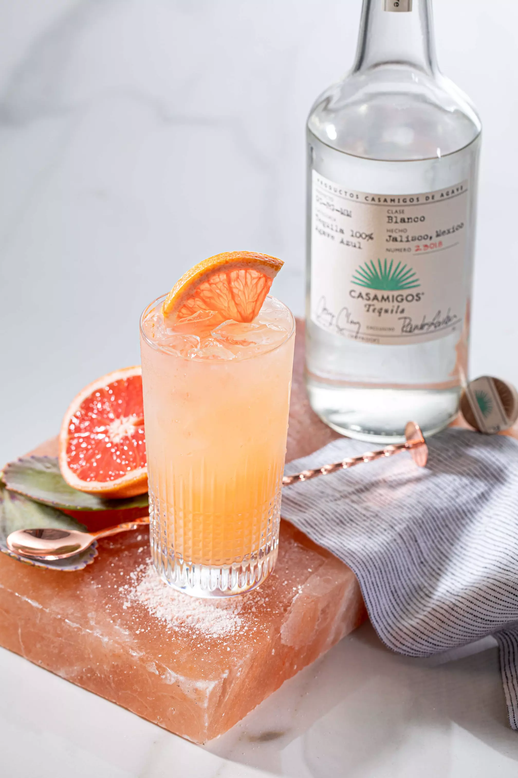 A tall glass of grapefruit cocktail garnished with a dried grapefruit slice sits on a pink salt block, next to a bottle of Casamigos tequila and cocktail utensils.