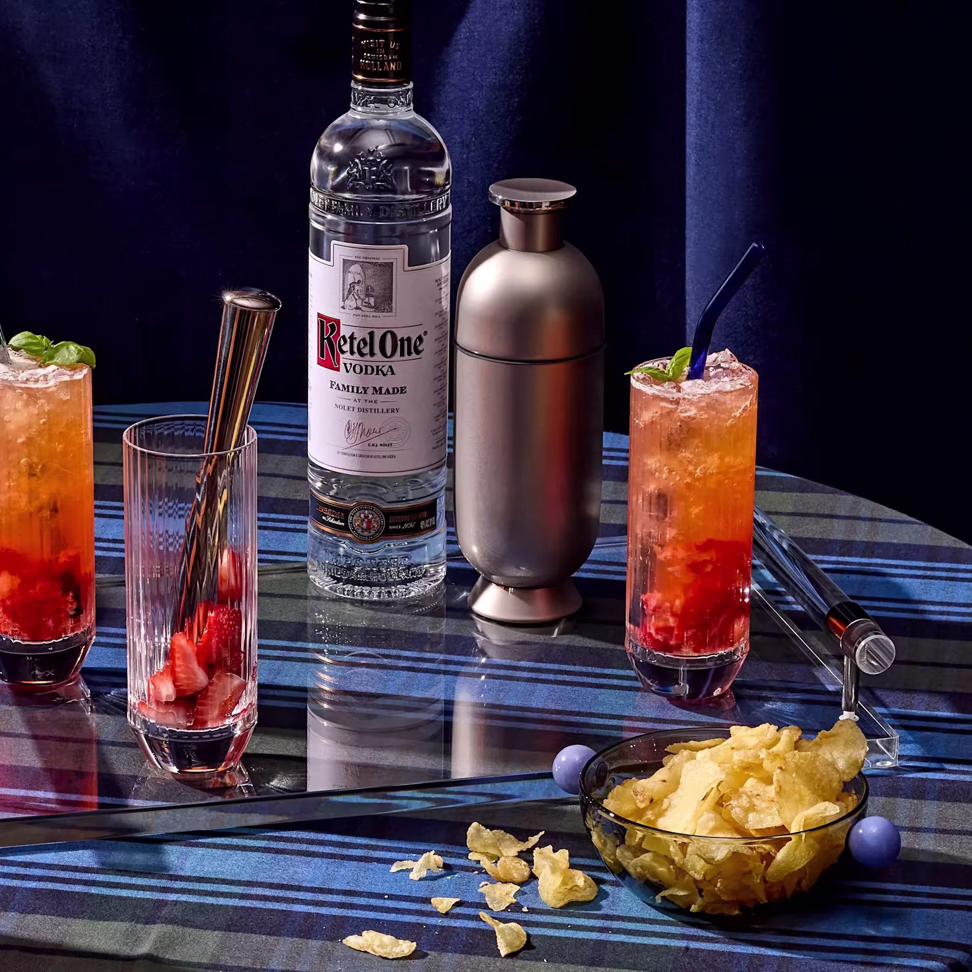 A bottle of Ketel One vodka, a cocktail shaker, two strawberry cocktails, a plate of potato chips, and straws are arranged on a blue plaid tablecloth.